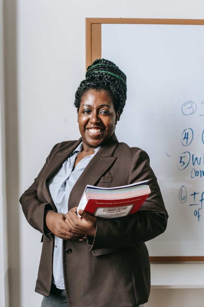 pexels-photo-5905483 happy black female teacher standing with workbooks near whiteboard