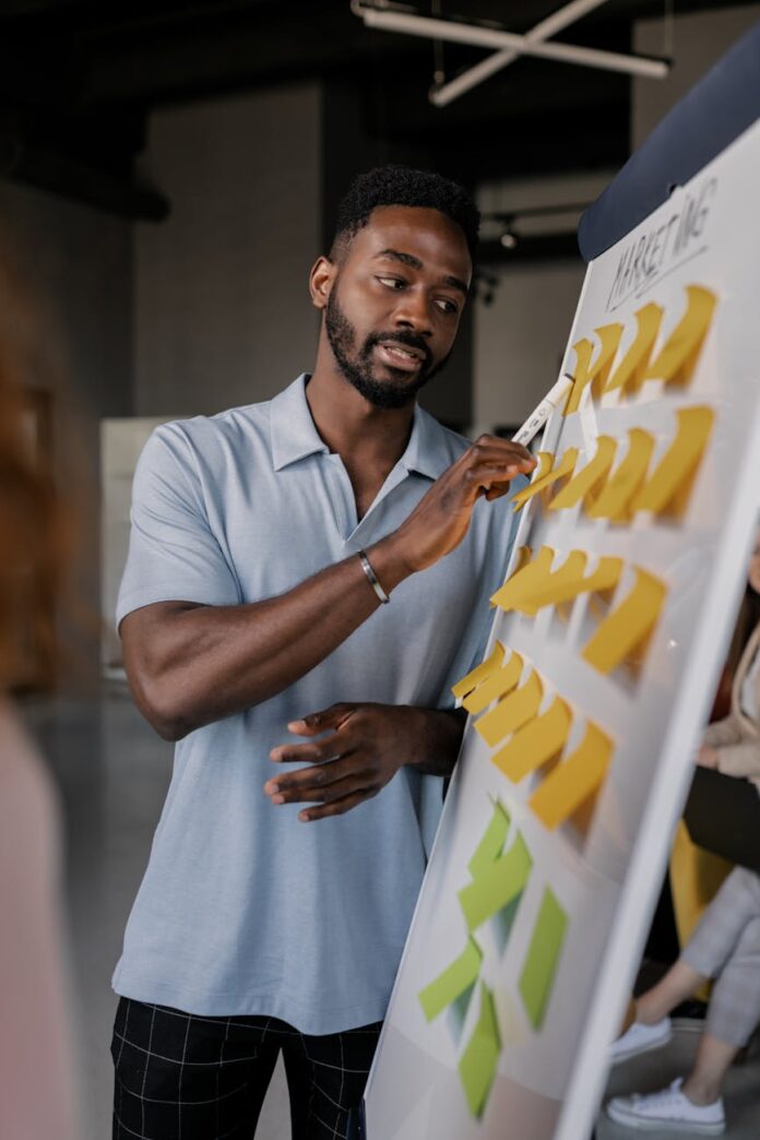pexels-photo-7710088 man in a blue shirt pointing to a whiteboard with sticky notes